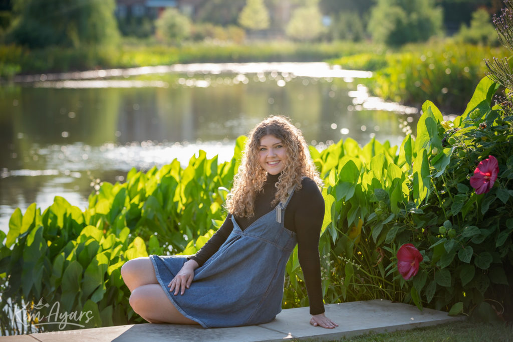 A high school senior is sitting on a concrete step in front of a pond with the sun behind her.