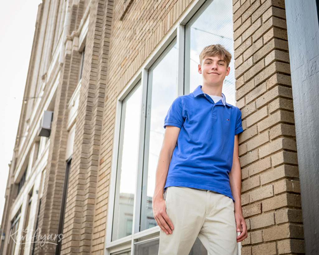 A high school senior in a blue shirt leans against a tall building in downtown Aurora.
