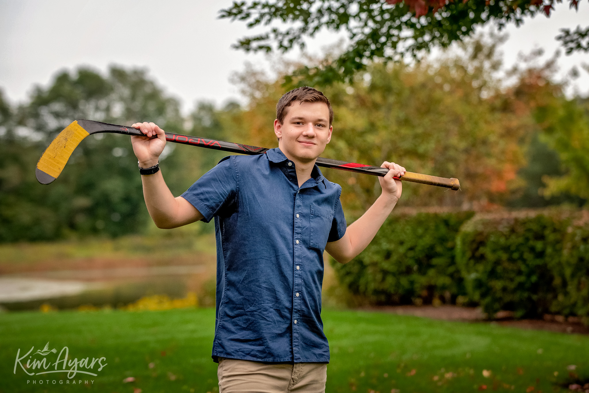 A high school senior is holding a hockey stick and smiling at the camera.