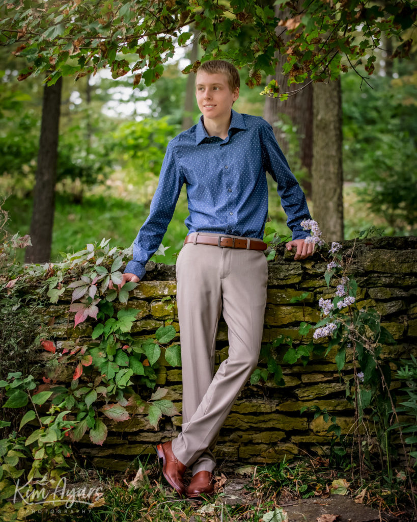 A high school senior is leaning against a stone wall in the springtime.
