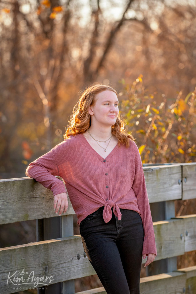 A high school senior is leaning against a bridge railing at sunset with fall foliage behind her.