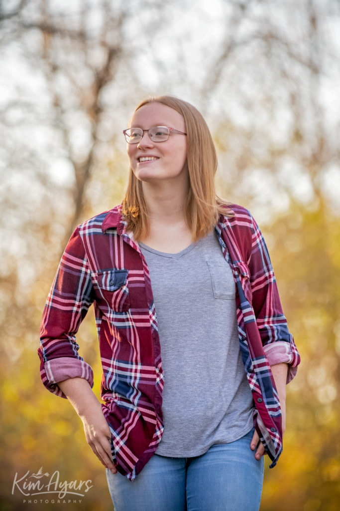 A high school senior in a plaid shirt is walking down a gravel path with golden colored leaves behind her at sunset.